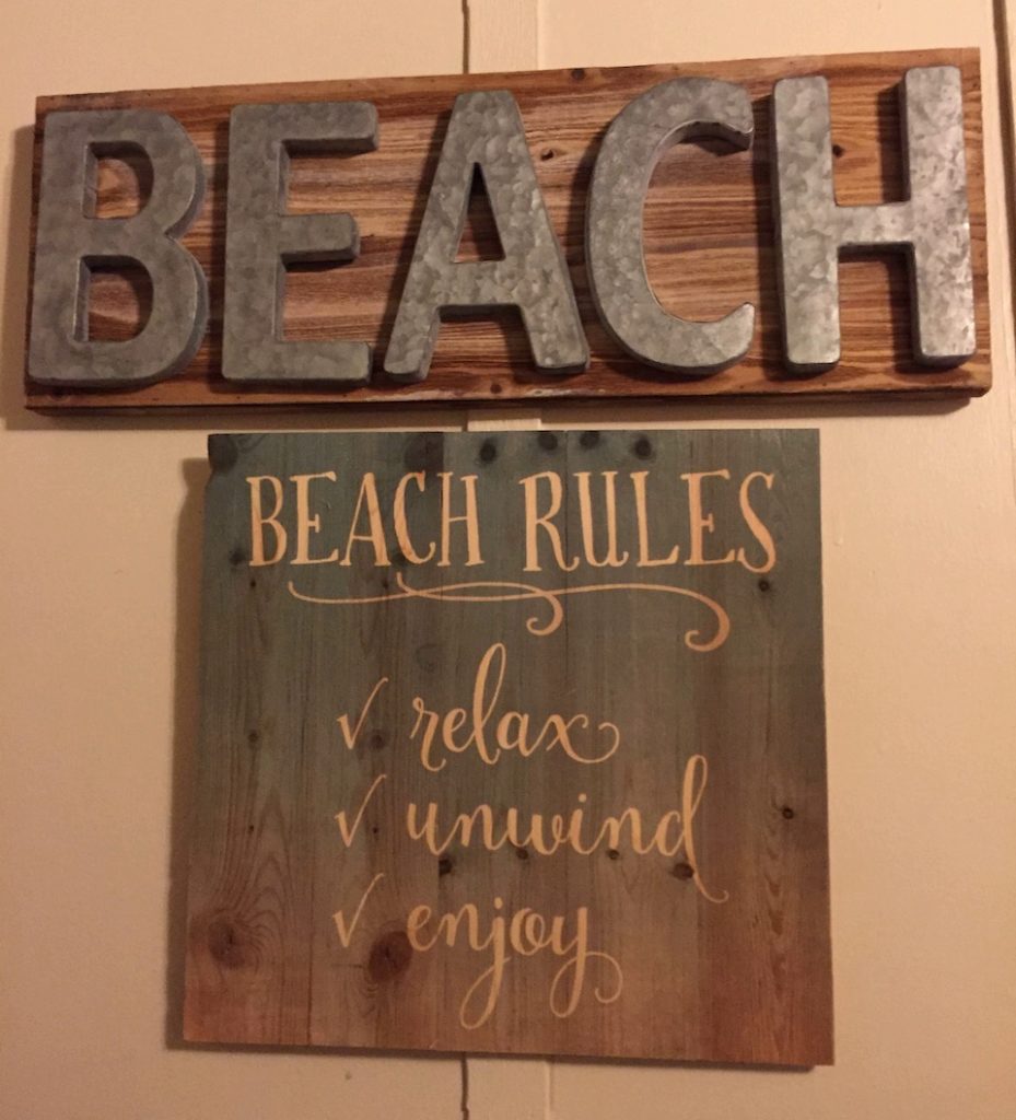 Two wooden plaques handing on a beige well. Top, rectangular plaque reads "BEACH." A square plaque beneath it reads "BEACH RULES: relax, unwind, enjoy."