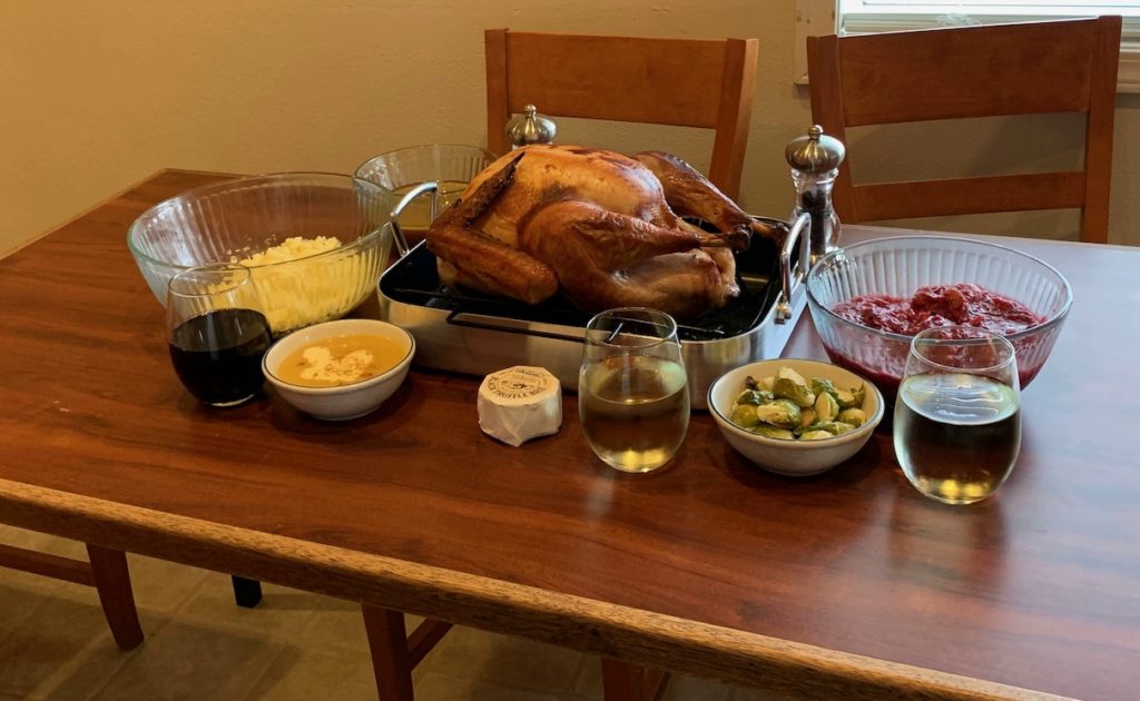 Dinner on the table: a roasted turkey in the middle, flanked by a bowl of mashed potatoes, a bowl of gravy and a glass of red wine on the left; two glasses of white wine, a bowl of brussels sprouts, and a bowl of cranberry compote on the right.