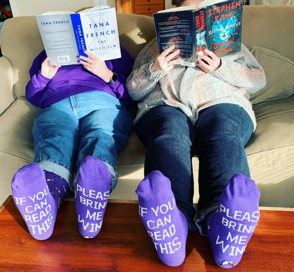 Two women relaxing on a couch, with their feet on the coffee table. Both are reading books (The Witch Elm by Tana French of the left, Sleeping Beauties by Stephen and Owen King on the right) that obscure their faces. They are both wearing socks that say "If you can read this" on one foot, "Please bring me wine" on the other.