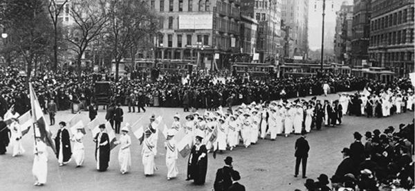 Historic photo: black and white image of a crowd of women suffragettes dressed in white marching on a city street lined by men in dark suits.