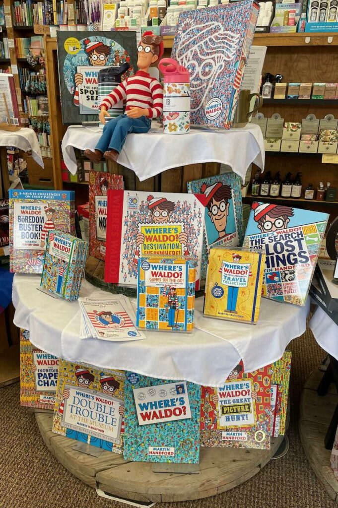 A 3-level round display featuring many "Where's Waldo?" books. On the top level sits a Waldo doll: A male figure with eyeglasses, wearing a red and while horizontal striped shirt and blue jeans.