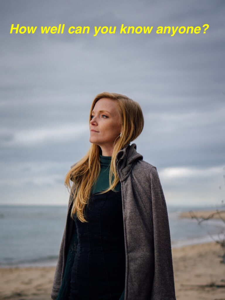 Photo. Background: slightly out-of-focus ocean and sandy beach. Foreground: Woman staring into distance off to left of picture.