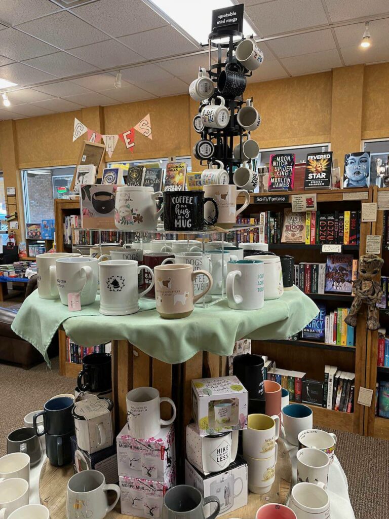 Background: bookshelves. Foreground: a 3-level round display featuring lots of mugs with book-related quotations.