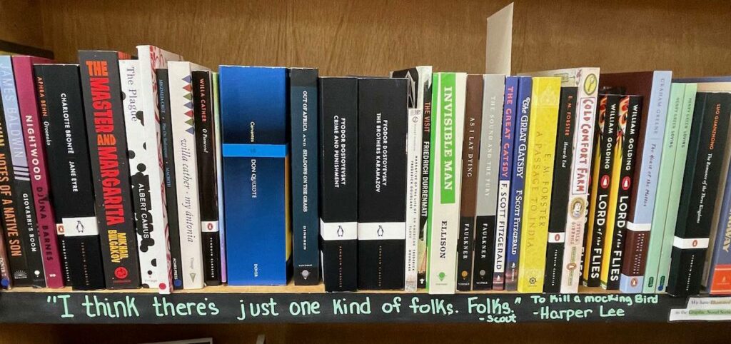 A long shelf of novels. Along the bottom edge of the shelf is a hand-lettered quotation: "I think there's just one kind of folks. Folks." Scout from To Kill a Mockingbird by Harper Lee