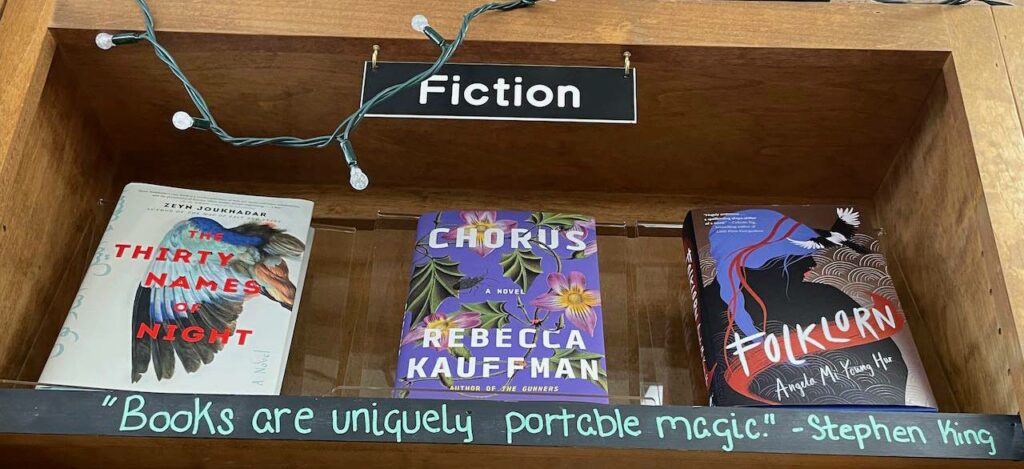 A shelf labeled "fiction" featuring 3 books: The Thirty Names of Night by Zeyn Joukhadar, Chorus by Rebecca Kauffman, and Folklorn by Angela Mi Young Hur. Along the bottom edge of the shelf is a hand-lettered quotation: "Books are Uniquely portable magic," Stephen King.