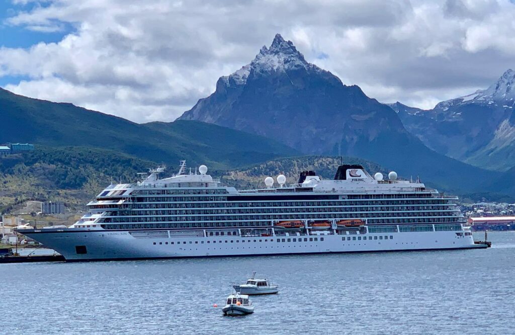 A small ocean cruise ship on the water, with green hills and a snow-covered mountain peak in the background.