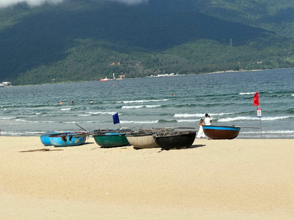 foreground: sandy beach with several open boats that resemble large, flat-bottom baskets. Background: ocean waves rolling toward beach.