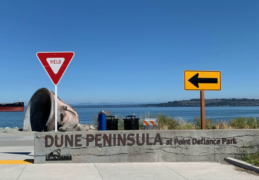 Foreground: Sign reading "Dune Peninsula at Point Defiance Park." Background: Blue sky over blue water, with mountains beyond.