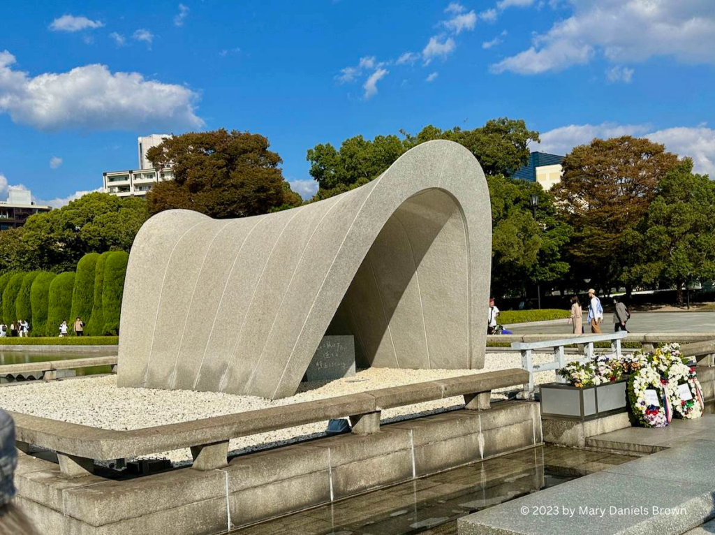 An arch-shaped monument covers a container containing the names of all victims of the atomic bomb dropped on Hiroshima, Japan, on August 6, 1945. To the right of the monument are flowers and wreaths of rememberance.