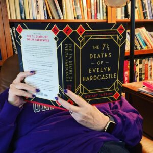 Woman in chair, with an open book held in front of her face. Book title: The 7 1/2 Deaths of Evelyn Hardcastle. Shelves in the background filled with books.