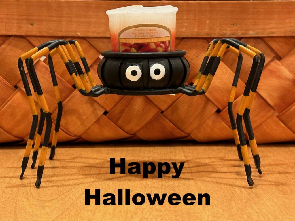 A small black open container, holding a white votive candle, with 4 orange-and-black bent legs on each side. Title: Happy Halloween