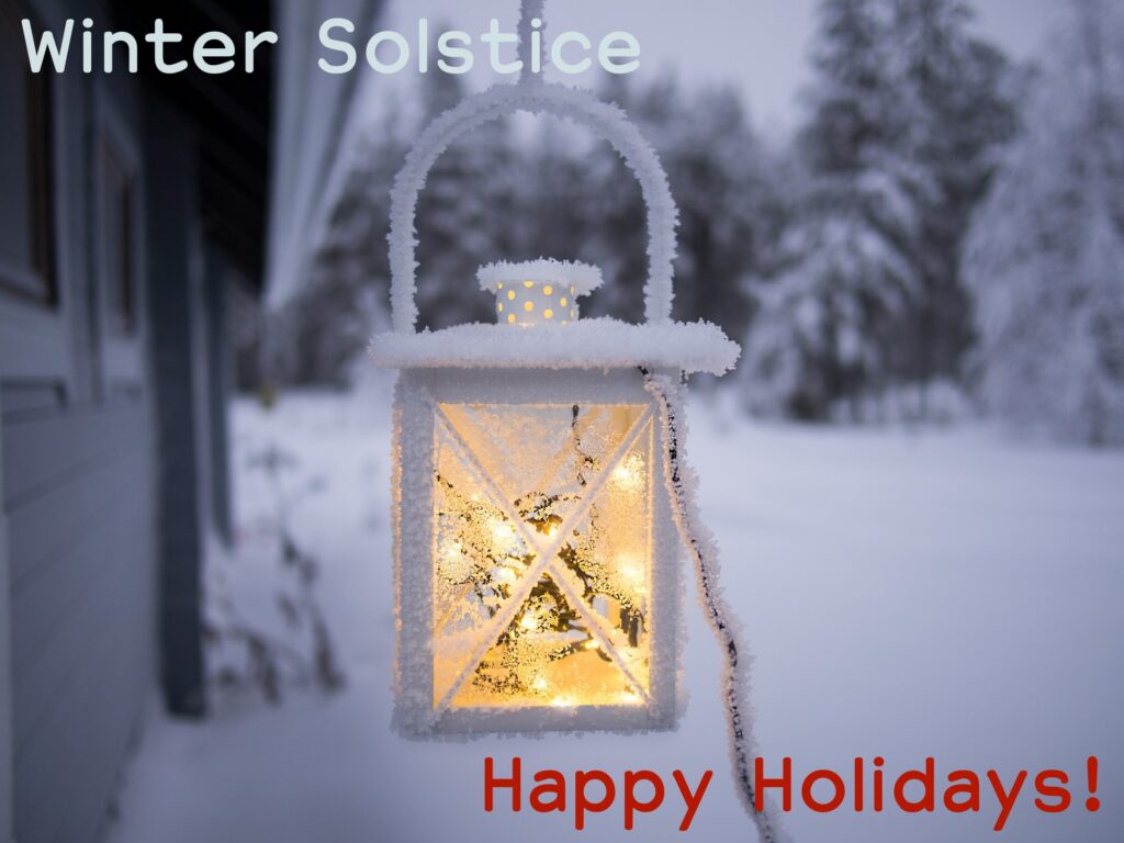 Background: slightly blurred view of snow-covered ground and trees. Foreground: Rectangular snow-covered lantern with soft light glowing inside.